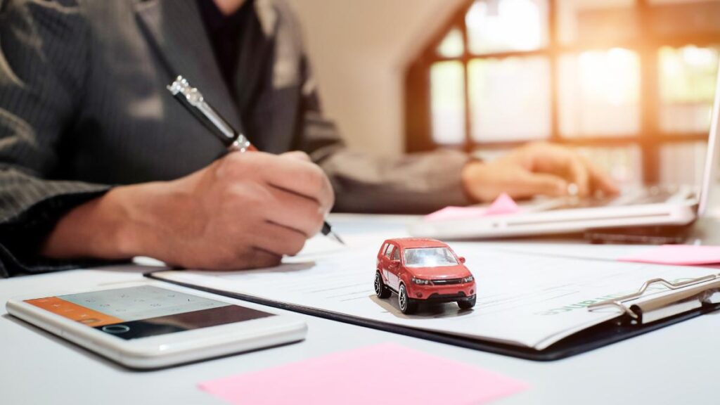 Homem de terno assinando contrato de seguro auto, com um carro vermelho em miniatura, calculadora e notebook sobre a mesa.
