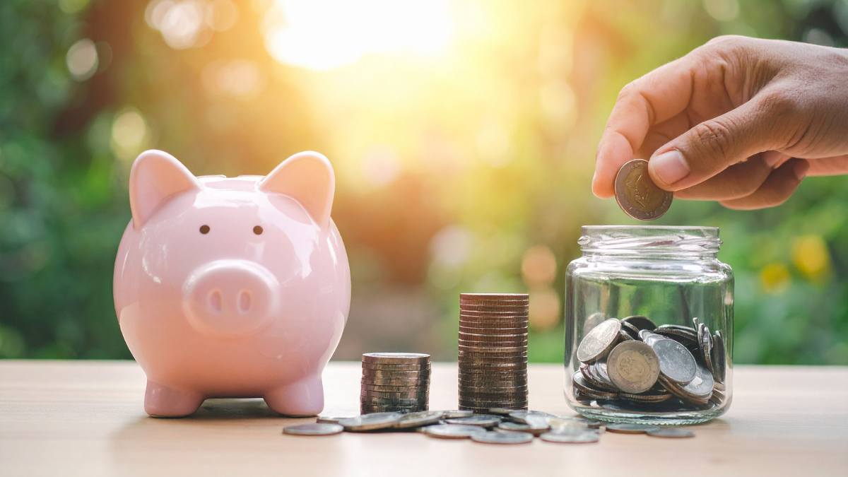 Mão colocando moeda em cofrinho de vidro ao lado de pilhas de moedas e porquinho rosa, ilustrando como economizar no seguro auto.