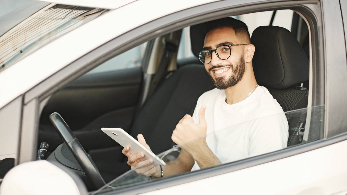 Homem sorridente dentro de um carro branco segurando um tablet, representando a conferência da renovação automática de seguro.