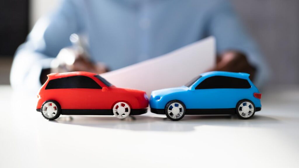 Dois carros de brinquedo colidindo sobre uma mesa com uma pessoa assinando contrato ao fundo, ilustrando a escolha entre seguro compreensivo ou terceiros.