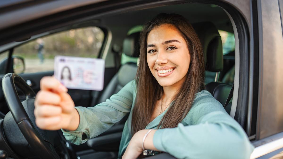 Mulher jovem e sorridente dentro de um carro segurando sua CNH, ilustrando o perfil de seguro para motorista iniciante.