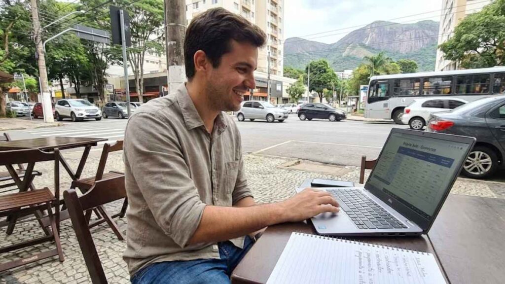 Homem sorrindo sentando em um café ao ar livre, usando um notebook para pesquisar formas de economizar no seguro auto em Belo Horizonte, com a serra ao fundo.