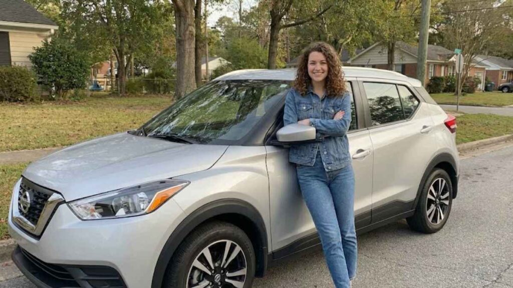 Moça jovem de 22 anos encostada em um Nissan Kicks prata em uma rua residencial, ilustrando o perfil de quem busca pelo Seguro Nissan Kicks para jovem.